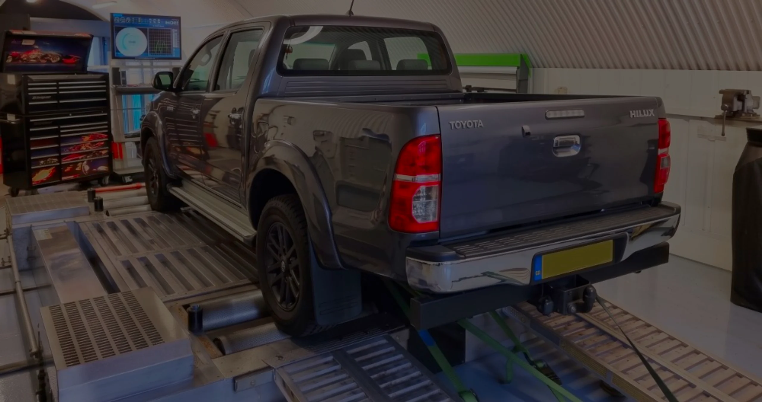 Dark grey Toyota Hilux rear three-quarter on rolling road dyno with HILUX tailgate lettering and green safety straps at 24Tuning Bournemouth Dorset workshop for Stage 1 ECU remapping