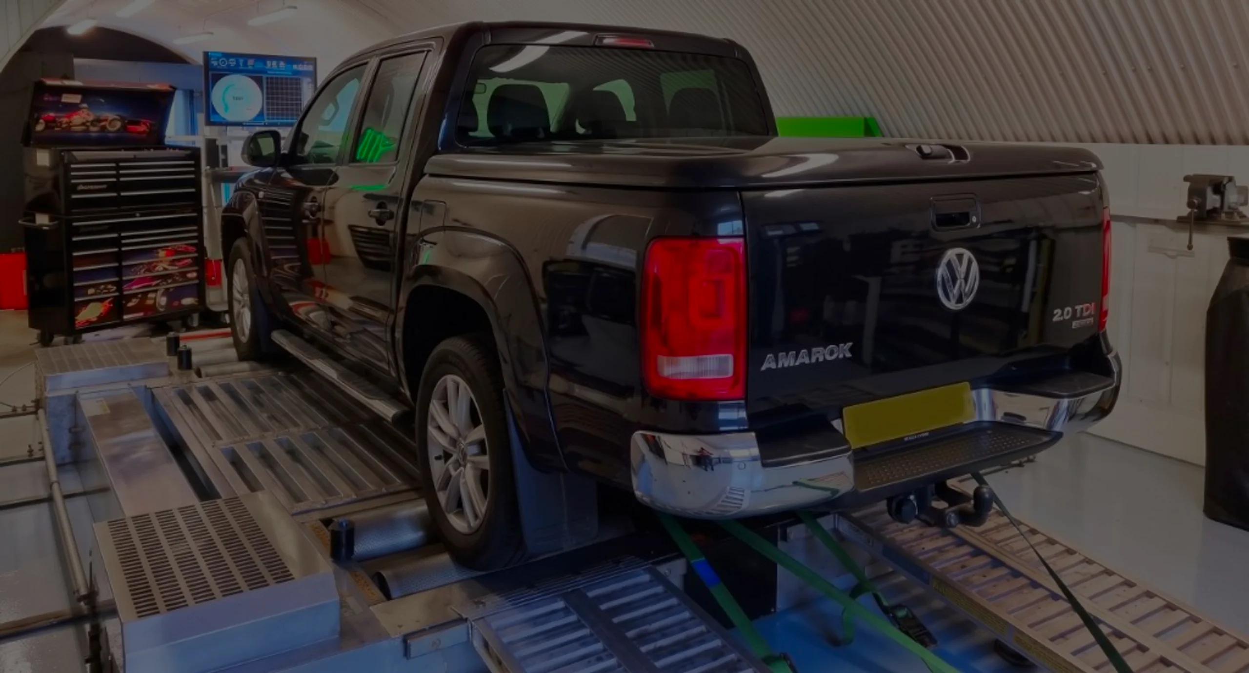 Black Volkswagen Amarok 2.0 TDI 4MOTION rear three-quarter on rolling road dyno with AMAROK tailgate lettering and green safety straps at 24Tuning Bournemouth Dorset workshop for Stage 1 ECU remapping
